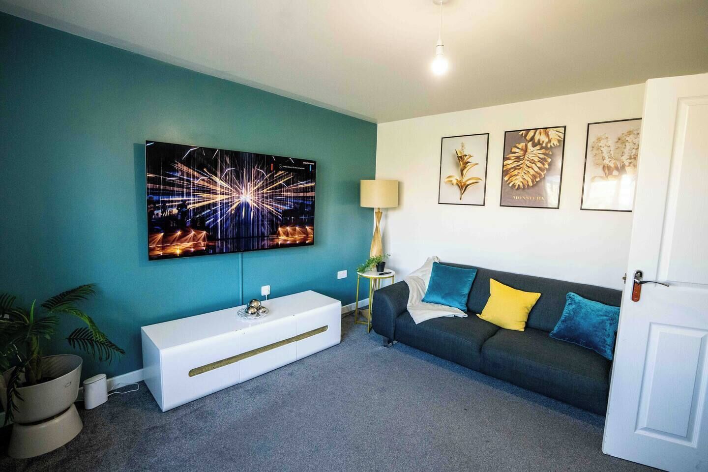 Ein Wohnzimmer mit türkisfarbenen Wänden, einem Flachbildfernseher, einer weißen Konsole und einem dunklen Sofa mit bunten Kissen sowie gerahmten Pflanzenbildern.