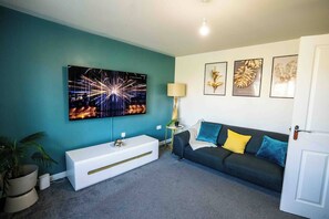 A living room with teal walls, a flat-screen TV, a white console table, a dark sofa with colorful cushions, and framed botanical prints. - 3-Bed Serviced Home | Ensuite | Work Desk| Bilston (West Midlands)