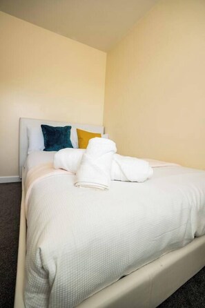 A neatly made bed with a white comforter and pillows, a headboard, and a plain wall. - 3-Bed Serviced Home | Ensuite | Work Desk| Bilston (West Midlands)