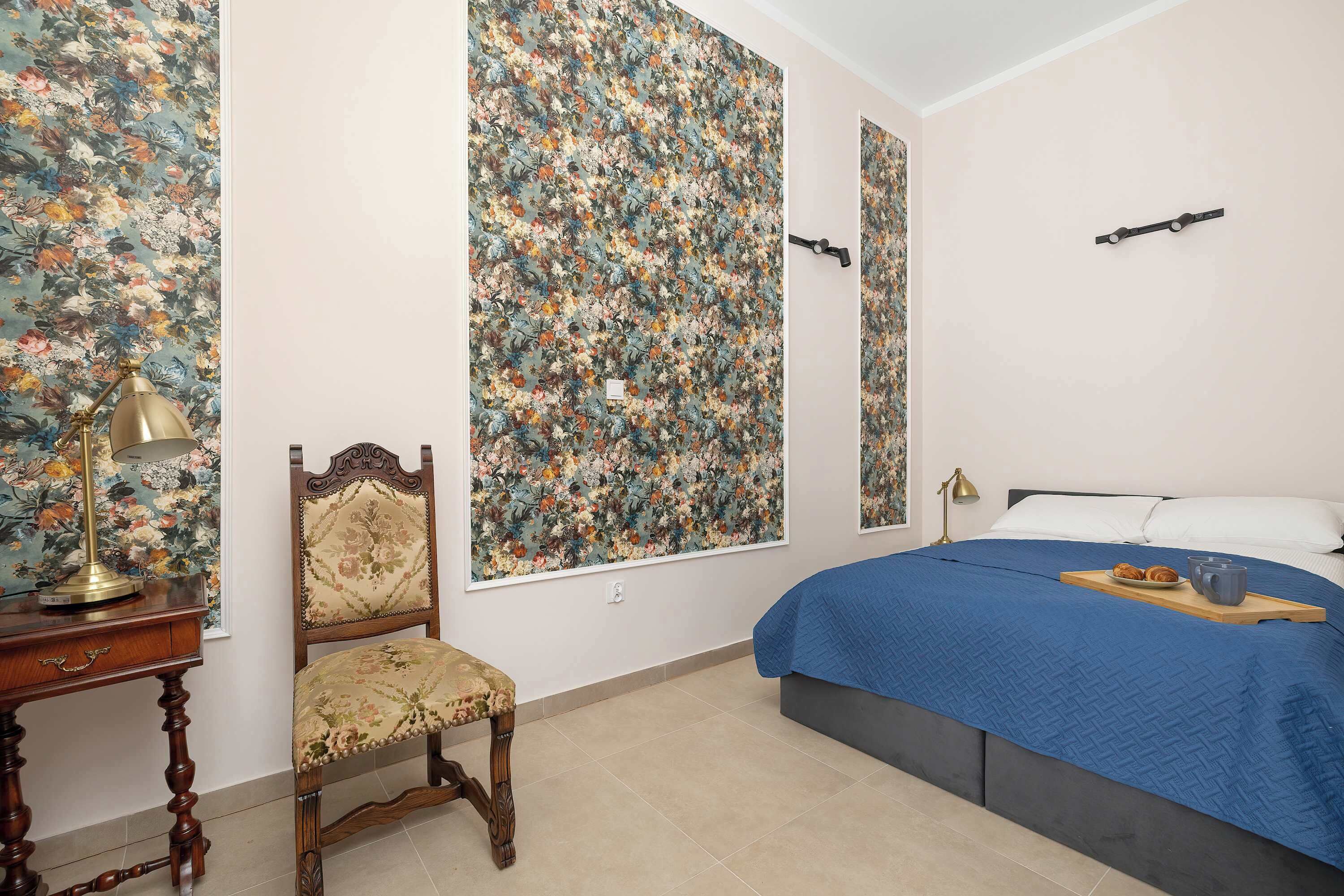 Ein Schlafzimmer mit einem Bett, einem Sessel, einem Schreibtisch und einer Wand mit floralem Tapetenmuster.