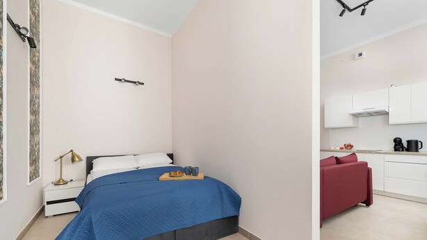A compact bedroom with a bed, bedside table, and a small desk with a lamp. Adjacent is a kitchenette with a red sofa, a coffee maker, and a kettle. - Charming Studio for 4 | Cosiness and Comfort (Bielsko-Biała)