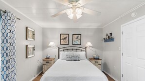 Un dormitorio con cama, dos mesitas de noche, ventilador de techo y cuadros en la pared.