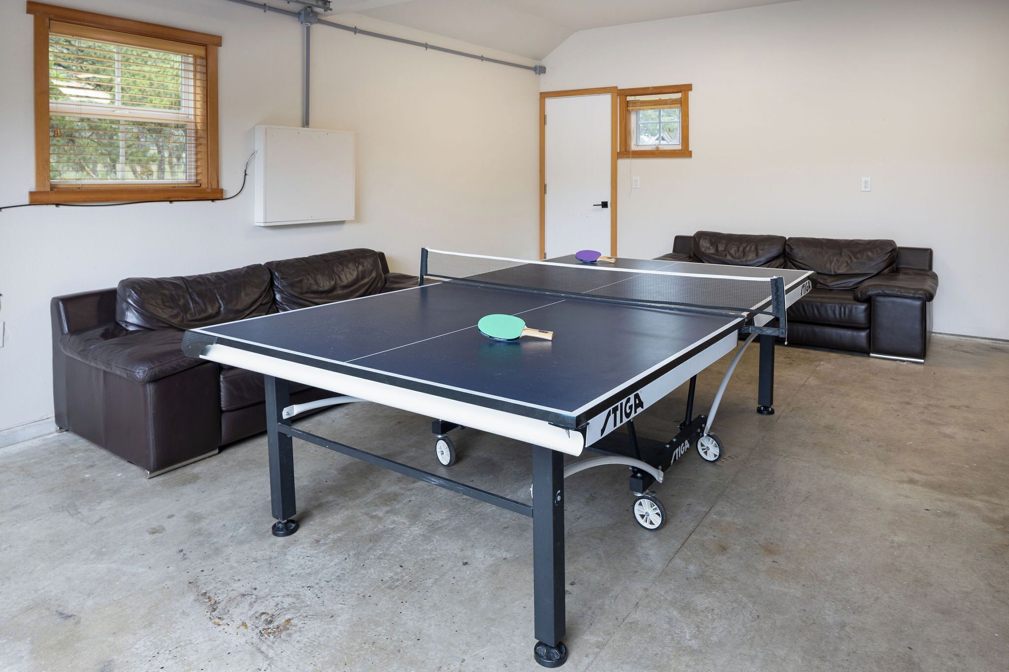Un tavolo da ping pong con ruote in una stanza con pavimento in cemento, due divani in pelle e una lavagna a parete.