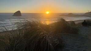 Een strand met een rotsformatie, gras en een persoon zittend in het zand.