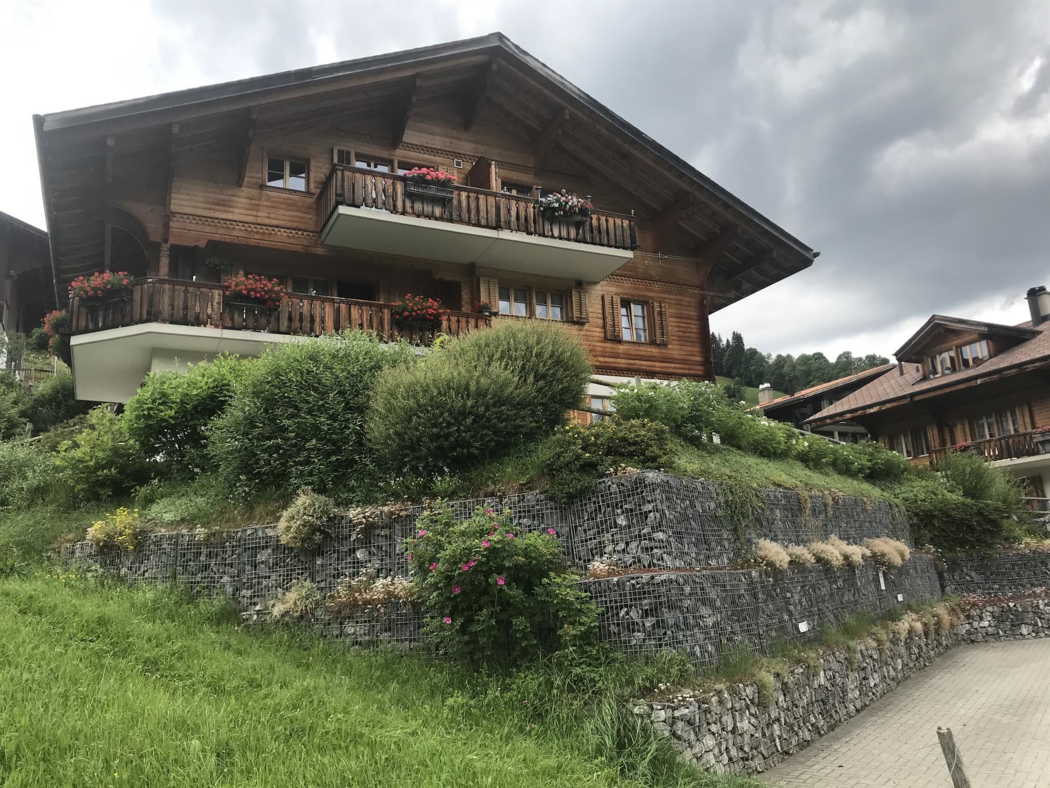 Un chalet en bois doté d’un balcon, entouré d’une végétation luxuriante et d’un mur de soutènement en pierre.