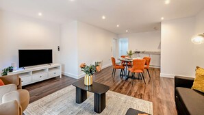 Uma sala de estar moderna com uma televisão de tela plana, uma área de refeições com cadeiras laranja e uma cozinha ao fundo.