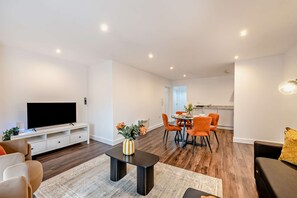 Ein modernes Wohnzimmer mit einem Flachbildfernseher, einem Essbereich mit orangefarbenen Stühlen und einer Küche im Hintergrund.