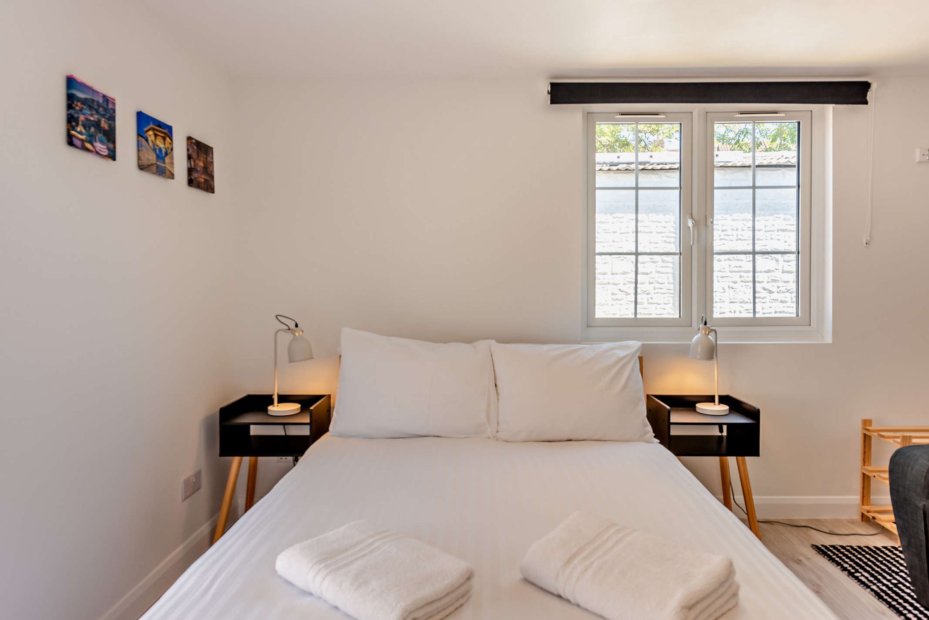 A bedroom with a bed, two bedside tables with lamps, a window with a view, and three wall-mounted pictures.