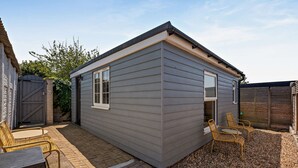 A small, single-story building with a grey exterior, a door, and a window. There are two wicker chairs and a small table outside. - Host Stay - Studio 58 (Brighton)