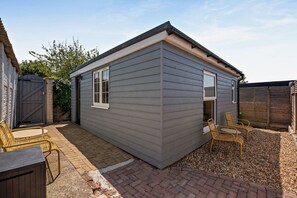 A small, single-story building with a grey exterior, a door, and a window. There are two wicker chairs and a small table outside. - Host Stay - Studio 58 (Brighton)