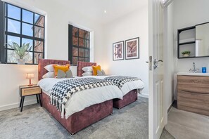 A bedroom with a large bed, two windows, a bedside table with a lamp, and a dresser with a mirror. - Stylish 2BR Apartment Jewellery Quarter 2 Ensuites Fast Wifi 8 Min to New St Station (Birmingham)