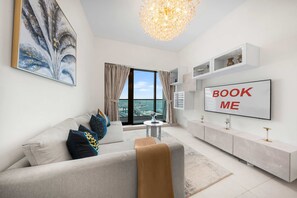 A modern living room with a sofa, a coffee table, a TV, and a view of the cityscape through large windows. - Free Housekeeping Visits During Your Stay - Stayshort - Modern 1BR in Prime Business Bay - 4 Guests (Dubai)