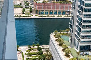 A high-rise building with a view of a body of water, a landscaped area with trees, and a commercial building with signage. - Free Housekeeping Visits During Your Stay - Stayshort - Modern 1BR in Prime Business Bay - 4 Guests (Dubai)