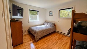 Ein Schlafzimmer mit Bett, Kommode, zwei Fenstern und einer Deckenleuchte.