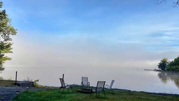 A lakeside view with chairs and a grill on the grass.