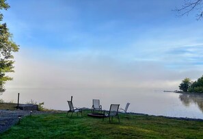 A lakeside view with chairs and a grill on the grass.
