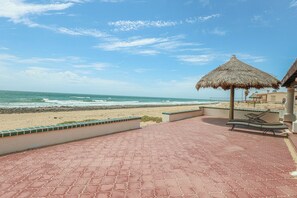 Eine Terrasse am Strand mit einem Reetdachschirm, Liegestühlen und Blick auf das Meer.
