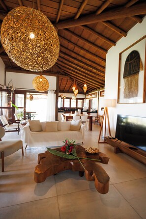 A spacious living area with a large woven light fixture, a wooden coffee table with a vase of flowers, a television, and a dining area in the background. - #casa_oceano_barra_grande Luxury Villa (MARAU)