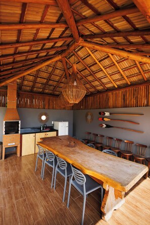 A rustic kitchen with a large wooden table, bar stools, a brick oven, and a hanging light fixture. - #casa_oceano_barra_grande Luxury Villa (MARAU)