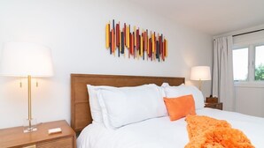 Een hotelkamer met een houten hoofdbord, witte beddengoed, een oranje plaid en een kunstwerk aan de muur.
