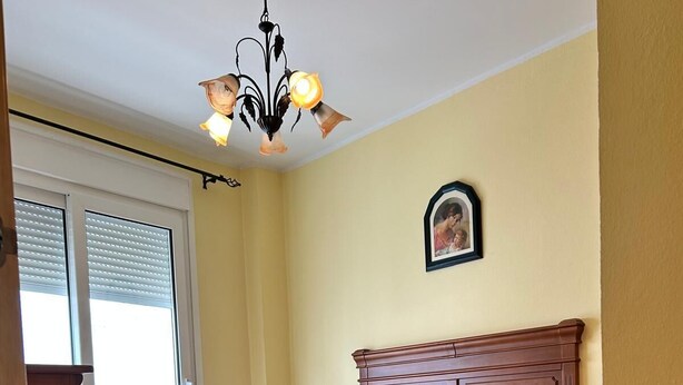 A bedroom with a bed, a nightstand, a wooden headboard, a chandelier, a window with blinds, and a framed picture on the wall. - Los Niños Apartment (Conil de la Frontera)