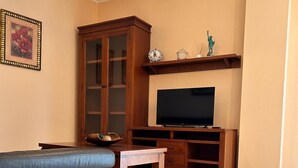 A living room with a sofa, a television on a wooden stand, a cabinet, and a wall-mounted shelf with decorative items. - Los Niños Apartment (Conil de la Frontera)