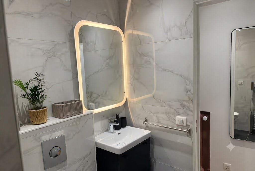 A modern bathroom with a marble wall, a white sink, a mirror with a light, and a potted plant.