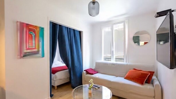 A modern living room with a sofa, a glass coffee table, a painting, a mirror, and a window with curtains.