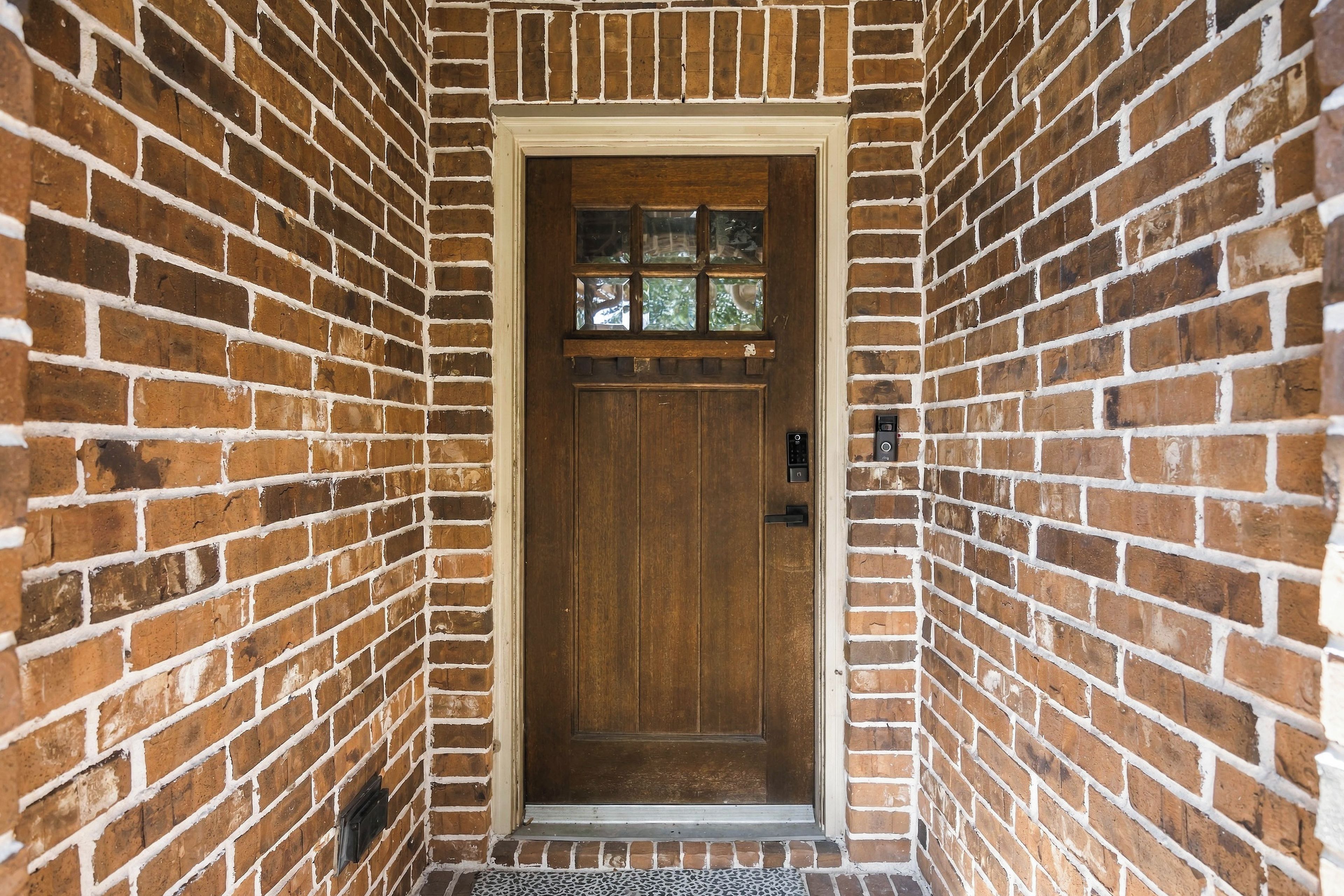 A brick entrance with a wooden door, a small window above the door, and a doormat.