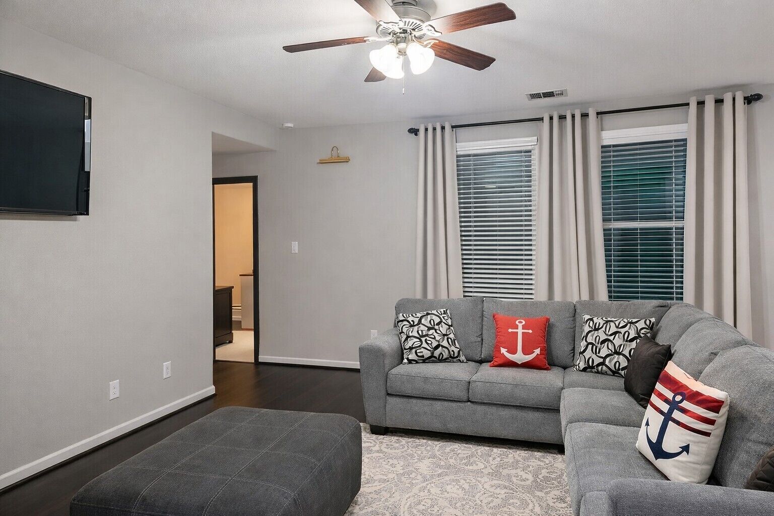 A living room with a grey sofa, ottoman, ceiling fan, and a flat-screen TV.