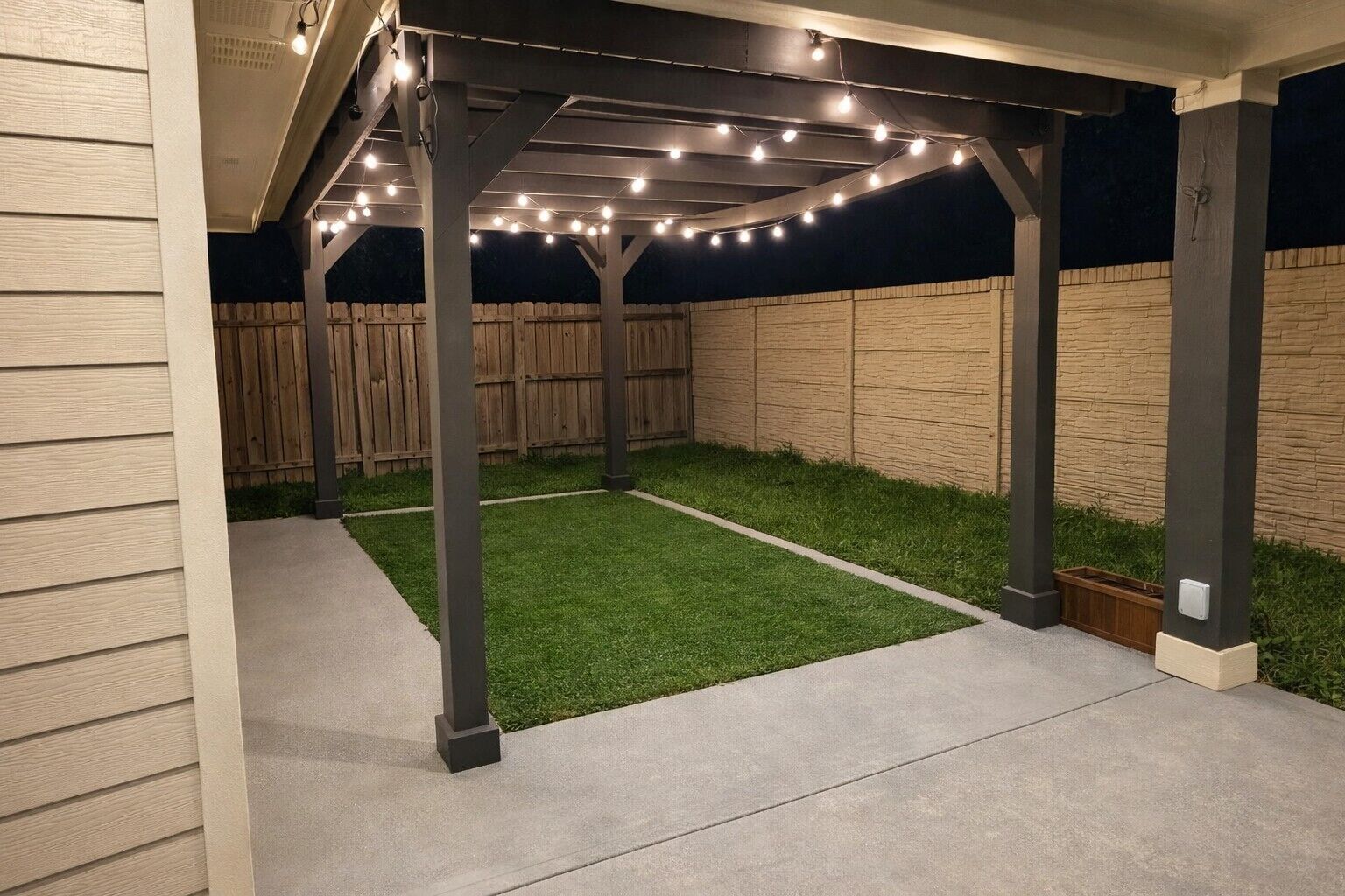 Een overdekt terras met kunstmatige verlichting, een betonnen vloer en een houten omheining op de achtergrond.
