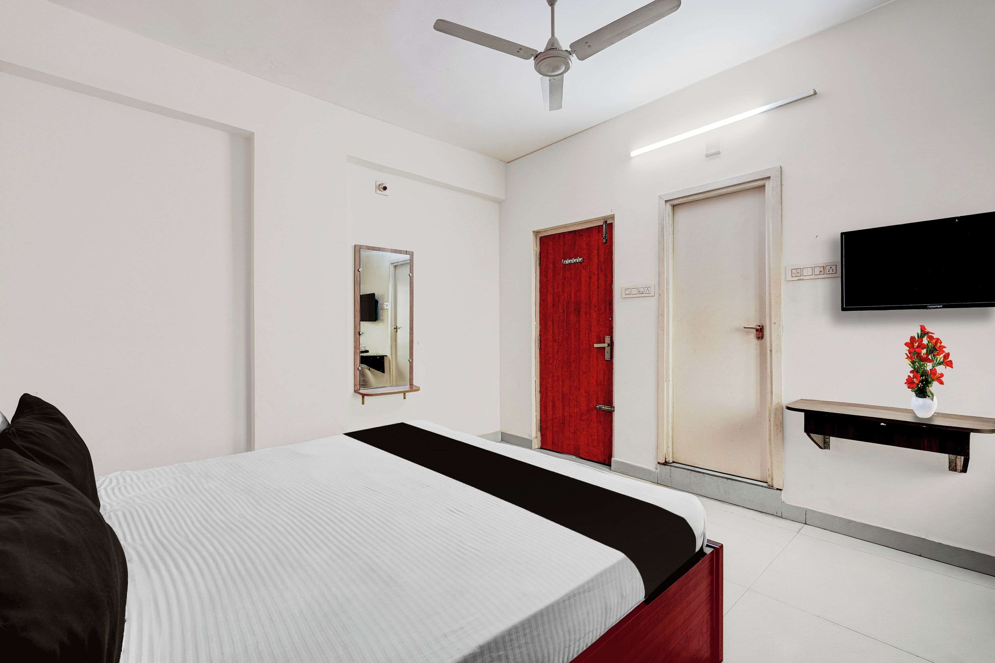 Habitación de hotel con cama, puerta roja, televisor de pantalla plana, espejo y una pequeña mesa con un florero.