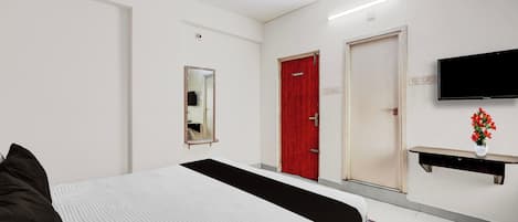 A hotel room with a bed, a red door, a flat-screen TV, a mirror, and a small table with a vase of flowers.
