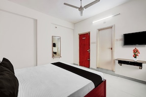 Habitación de hotel con cama, puerta roja, televisor de pantalla plana, espejo y una pequeña mesa con un florero.