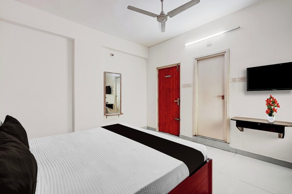 A hotel room with a bed, a red door, a flat-screen TV, a mirror, and a small table with a vase of flowers.