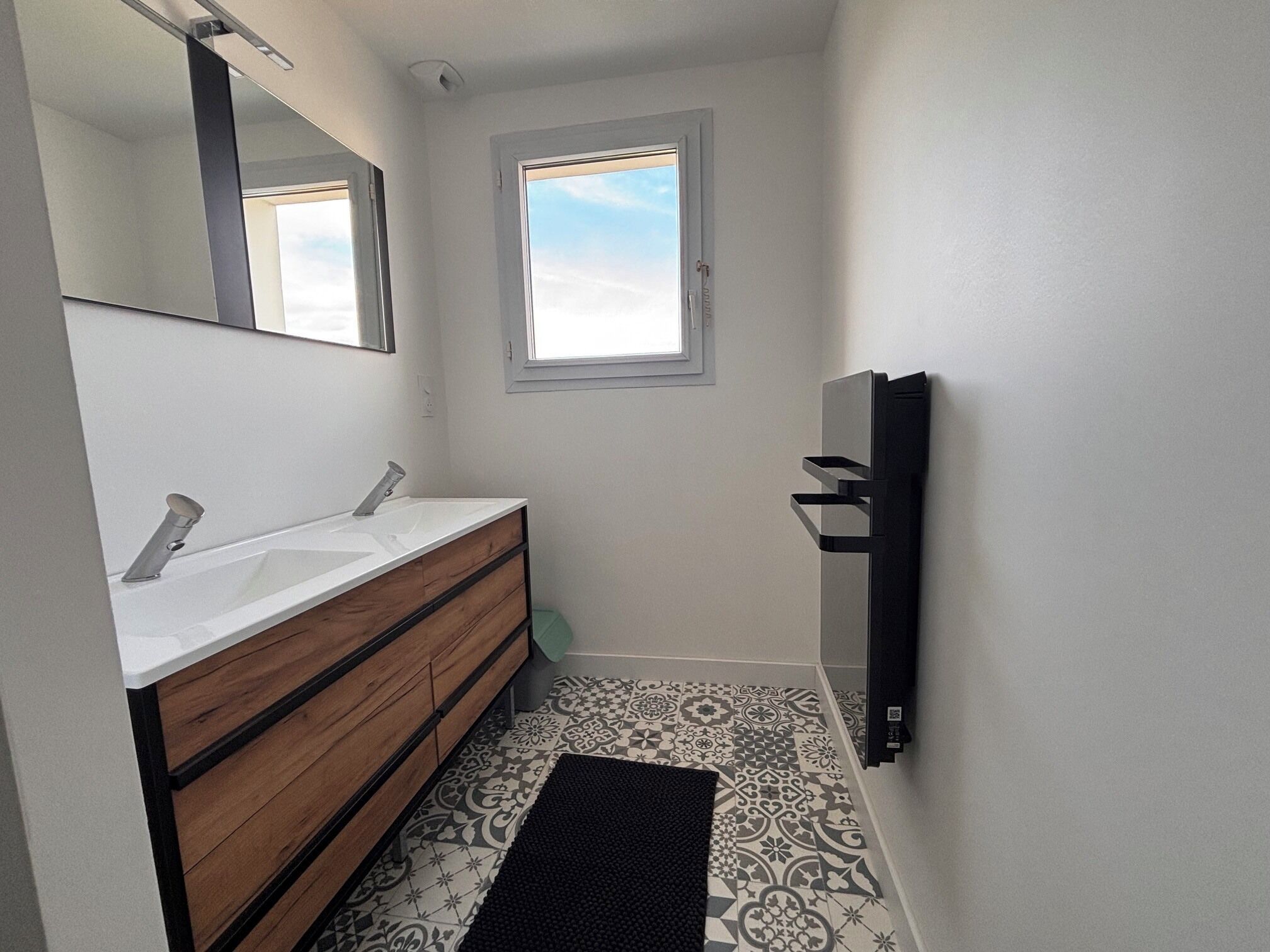 Ein Badezimmer mit doppeltem Waschbecken, Holzschrank, gemusterten Bodenfliesen, einer schwarzen Matte, einem Fenster und einem Handtuchhalter an der Wand.
