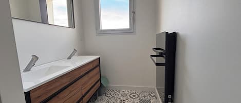 A bathroom with a double sink, wooden vanity, patterned floor tiles, a black mat, a window, and a wall-mounted towel rack.