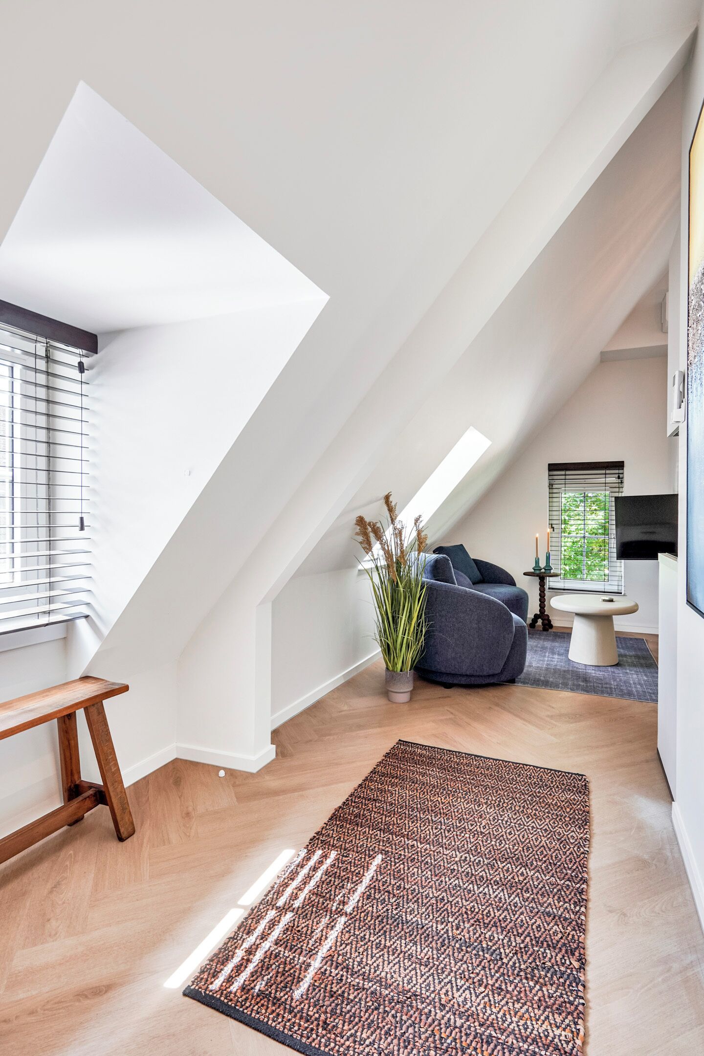 A modern loft bedroom with a sloped ceiling, a large skylight, a cozy seating area with a sofa and a small table, and a decorative plant.
