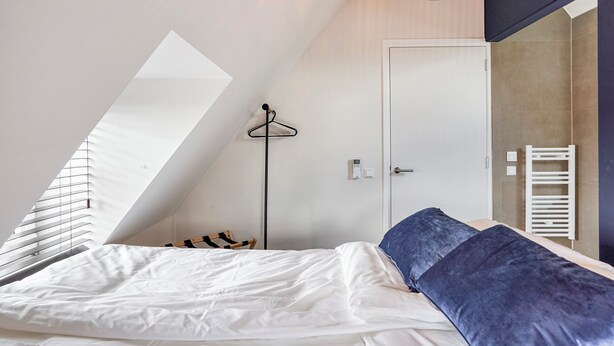 A bedroom with a sloped ceiling, a bed with white and blue bedding, a hanging light fixture, a coat rack, and a door with a towel rack. - Enjoy a stylish 50m² apartment for two in the heart of Velden. Featuring a comfortable queen bed, private kitchen, Smart TV, air conditioning, fast WiFi, and a shared balcony, it's an ideal base for both leisure and business stays. (Velden)