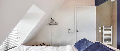 A bedroom with a sloped ceiling, a bed with white and blue bedding, a hanging light fixture, a coat rack, and a door with a towel rack.