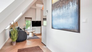 A modern living space with a high ceiling, a flat-screen TV, a grey sofa, and a large abstract painting. - Enjoy a stylish 50m² apartment for two in the heart of Velden. Featuring a comfortable queen bed, private kitchen, Smart TV, air conditioning, fast WiFi, and a shared balcony, it's an ideal base for both leisure and business stays. (Velden)