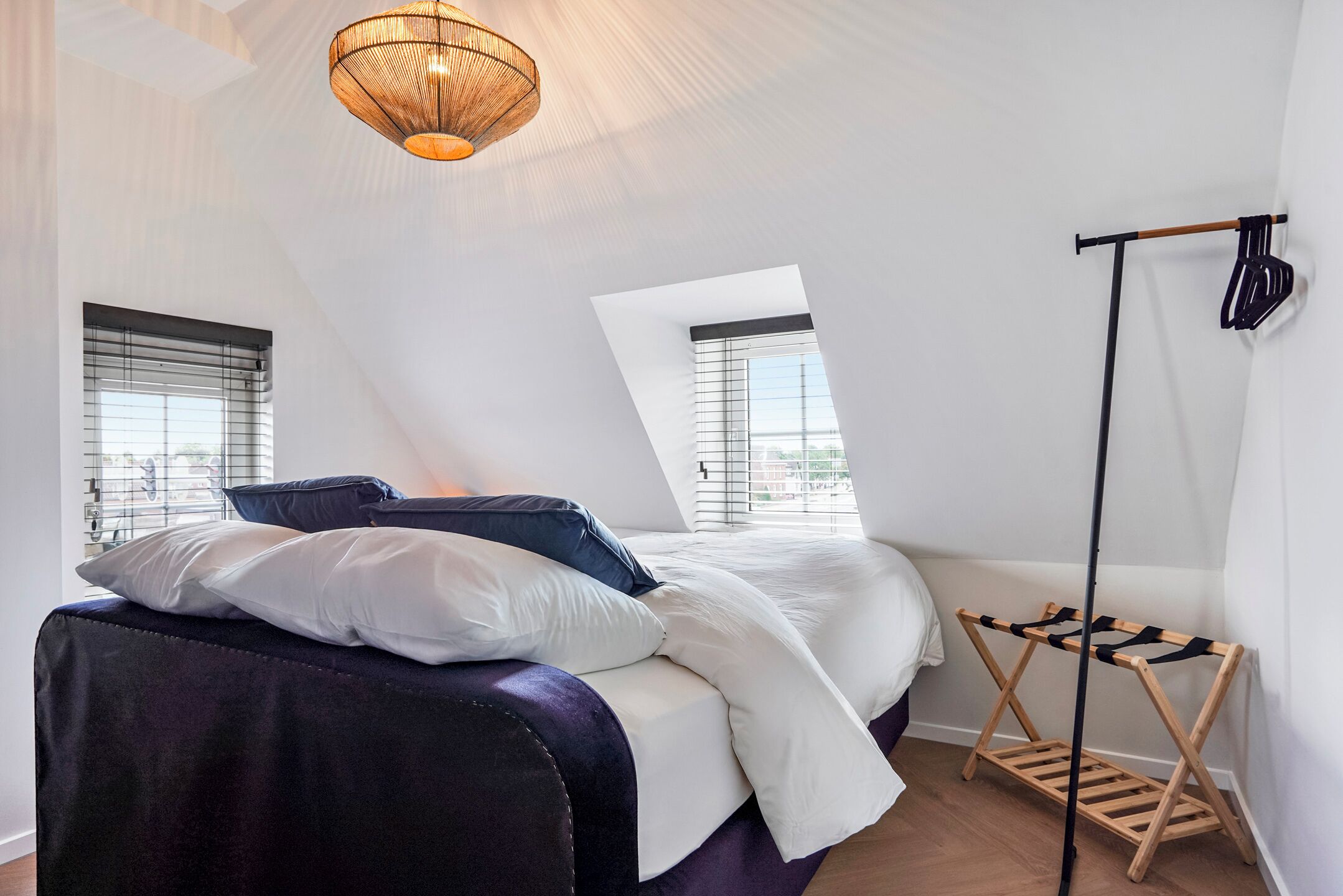 A bedroom with a sloped ceiling, a bed with white and dark bedding, a wooden drying rack, and a hanging light fixture.