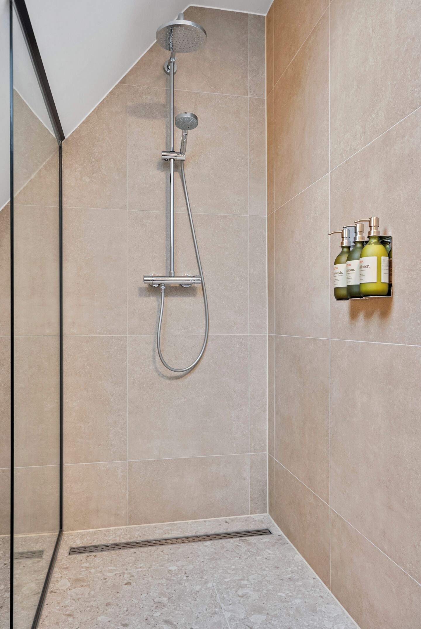 Eine Dusche mit Handbrause und Wandarmatur, ein gefliestes Badezimmer und ein Ablagefach mit Shampoo- und Conditionerflaschen.