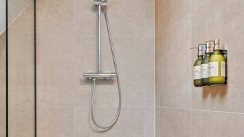 Eine Dusche mit Handbrause und Wandarmatur, ein gefliestes Badezimmer und ein Ablagefach mit Shampoo- und Conditionerflaschen.