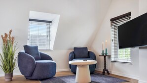 A modern living room with two blue armchairs, a round table, a potted plant, and a flat-screen TV mounted on the wall. - Enjoy a stylish 50m² apartment for two in the heart of Velden. Featuring a comfortable queen bed, private kitchen, Smart TV, air conditioning, fast WiFi, and a shared balcony, it's an ideal base for both leisure and business stays. (Velden)