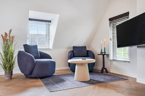 A modern living room with two blue armchairs, a round table, a potted plant, and a flat-screen TV mounted on the wall. - Enjoy a stylish 50m² apartment for two in the heart of Velden. Featuring a comfortable queen bed, private kitchen, Smart TV, air conditioning, fast WiFi, and a shared balcony, it's an ideal base for both leisure and business stays. (Velden)