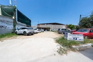 Un parking rempli de voitures, un panneau indiquant «Parking du véritable restaurant grec», et un bâtiment dont une partie est recouverte d’une bâche verte.