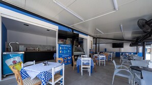 En restaurantindretning med skotske ternborde, hvide stole, et blåt og hvidt farvetema, et køkkenområde og et skilt, der reklamerer for cocktails og desserter.
