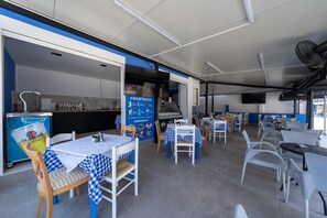 A restaurant interior with checkered tablecloths, white chairs, a blue and white color scheme, a kitchen area, and a sign advertising cocktails and desserts.