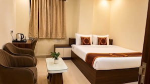 A hotel room with a bed, two chairs, a small table with a plant, a bedside table with a kettle, and a window with curtains.